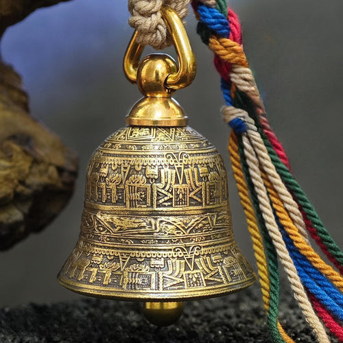"An antiqued brass Tibetan-style prayer bell, intricately engraved with traditional Buddhist scriptures and symbolic motifs across its curved surface. It is suspended from a polished gold-tone metal ring attached to a braided natural fiber cord, paired with vibrant, multicolored braided tassels in red, blue, green, yellow, cream, and brown. The bell rests against a weathered, dark stone formation against a moody, blurred gray-blue backdrop."