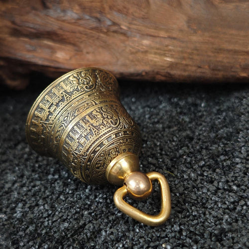 "An antiqued brass Tibetan-style prayer bell pendant, intricately engraved with traditional Buddhist scriptures, symbolic motifs, and ornate scrollwork across its entire curved surface. It is topped with a polished gold-tone metal bail and ring for hanging, resting on a dark, textured volcanic rock surface, with weathered driftwood in the background."