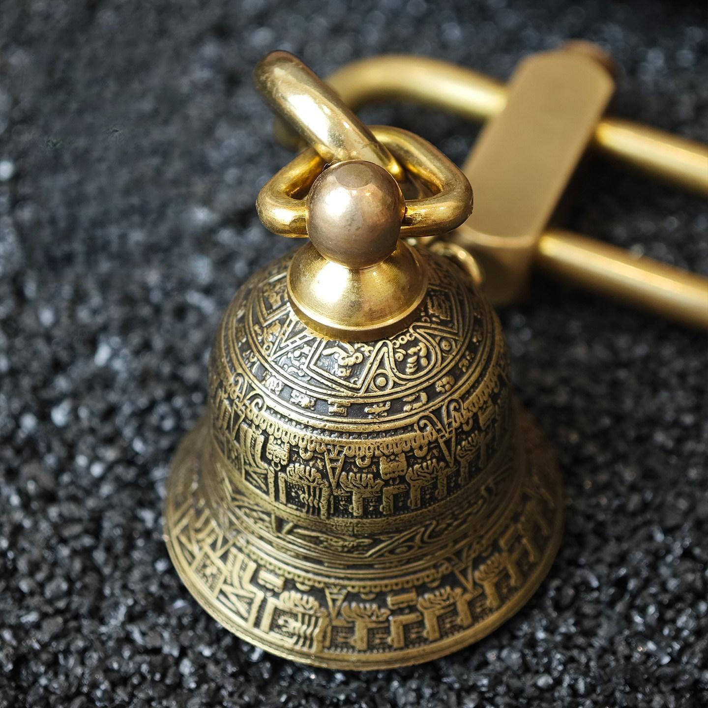 "An antiqued brass Tibetan-style prayer bell pendant, adorned with intricate engravings of traditional Buddhist scriptures and symbolic motifs across its entire surface. It features a polished gold-tone bail and ring for hanging, resting on a dark, textured asphalt surface next to a sleek, gold-tone metal belt buckle."