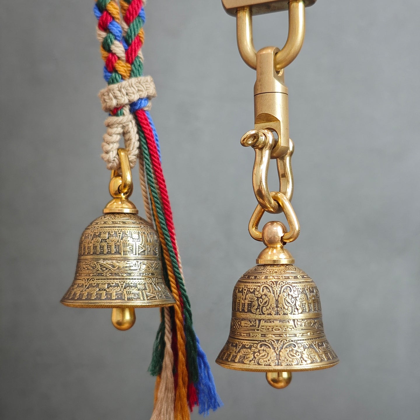 "A pair of antiqued brass Tibetan-style prayer bell pendants, both intricately engraved with traditional Buddhist scriptures and symbolic motifs across their curved surfaces. The left bell hangs from a vibrant, multicolored braided cord (featuring red, blue, green, yellow, and natural beige threads) secured with a hand-knotted fiber wrap and finished with a fringed tassel. The right bell is suspended from a polished gold-tone metal chain and heavy-duty clasp. The arrangement is set against a plain, muted gr