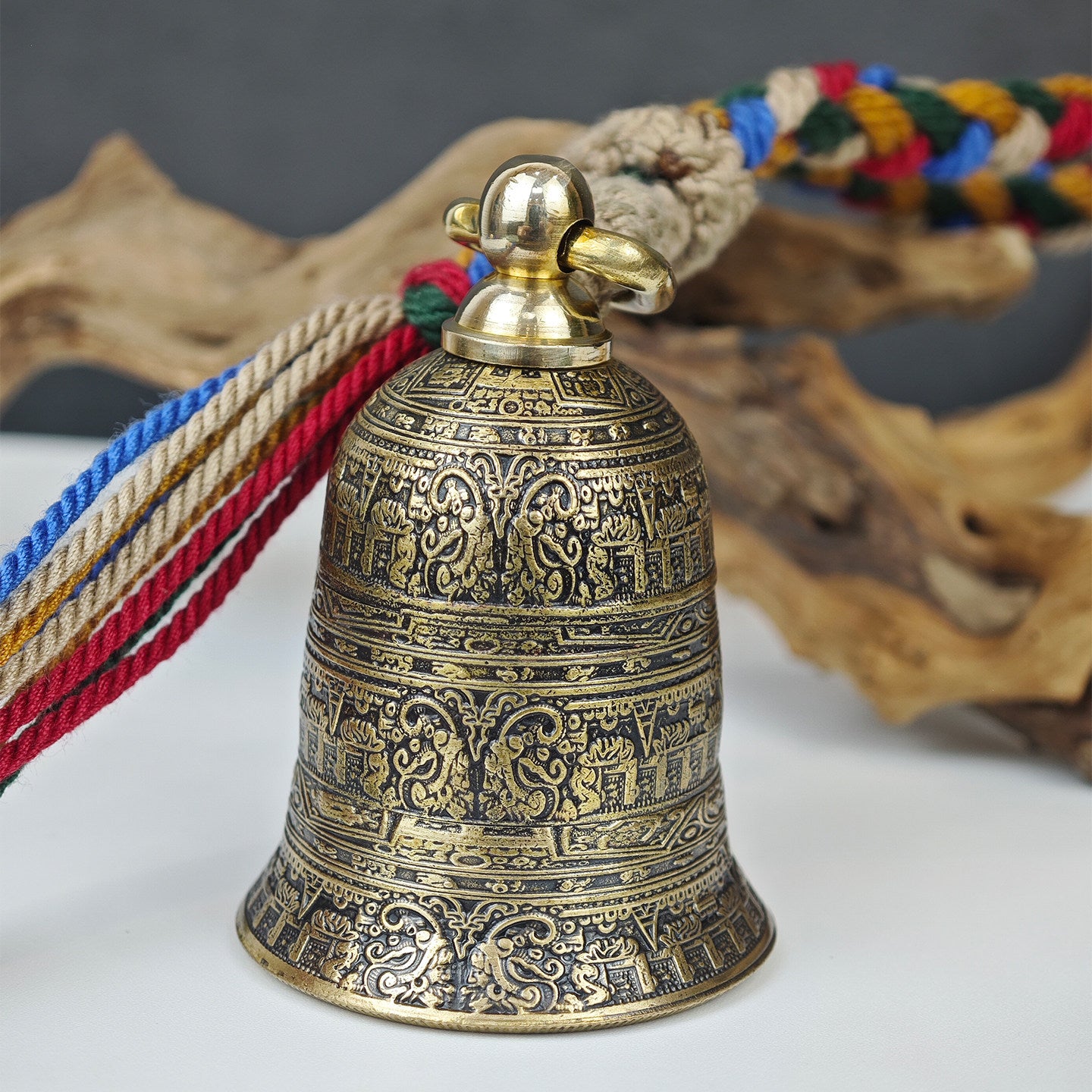 "An antiqued brass Tibetan-style prayer bell pendant, meticulously engraved with intricate bands of traditional Buddhist scriptures, symbolic figures, and ornate scrollwork across its entire curved surface. It is topped with a polished gold-tone metal bail and ring, attached to a vibrant, multicolored braided cord (featuring red, blue, green, yellow, and natural beige threads) finished with a fringed tassel. The bell rests on a clean white surface, with weathered, gnarled driftwood and a muted gray backdrop