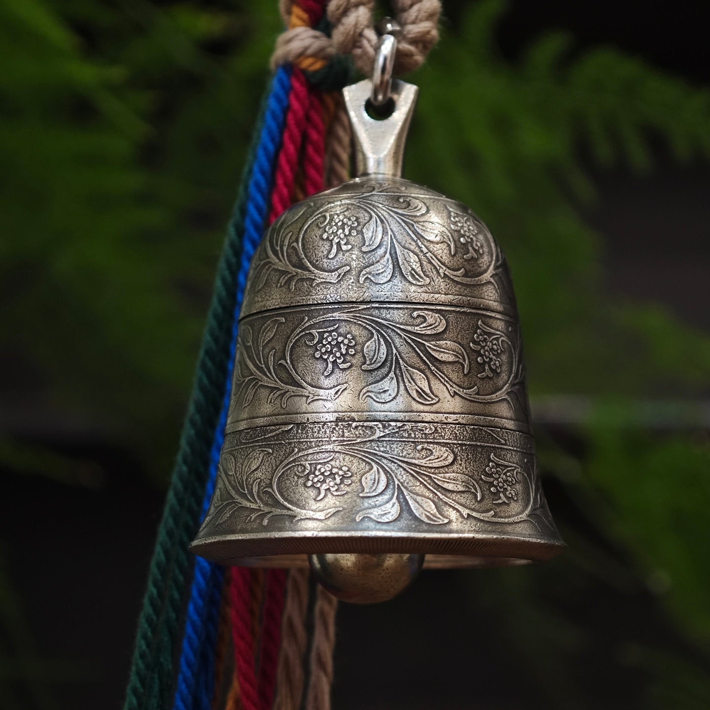 "An ornate, antiqued silver-toned metal bell pendant, featuring three distinct horizontal bands of intricate hand-engraved floral scrollwork and small flower clusters across its curved surface. A smooth metal clapper is visible inside the bell’s opening. It is suspended from a polished silver-toned metal bail and jump ring, paired with a collection of vibrant, multicolored braided cords (featuring green, blue, red, and natural beige threads) twisted together and secured with a woven fiber wrap at the top. T