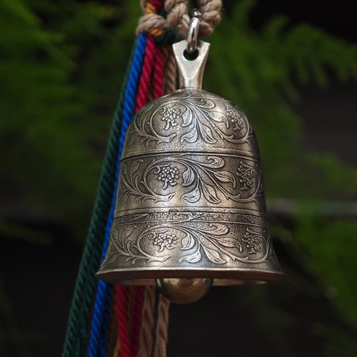 "An ornate, antiqued silver-toned metal bell pendant, featuring three distinct horizontal bands of intricate hand-engraved floral scrollwork and small flower clusters across its curved surface. A smooth metal clapper is visible inside the bell’s opening. It is suspended from a polished silver-toned metal bail and jump ring, paired with a collection of vibrant, multicolored braided cords (featuring green, blue, red, and natural beige threads) twisted together and secured with a woven fiber wrap at the top. T
