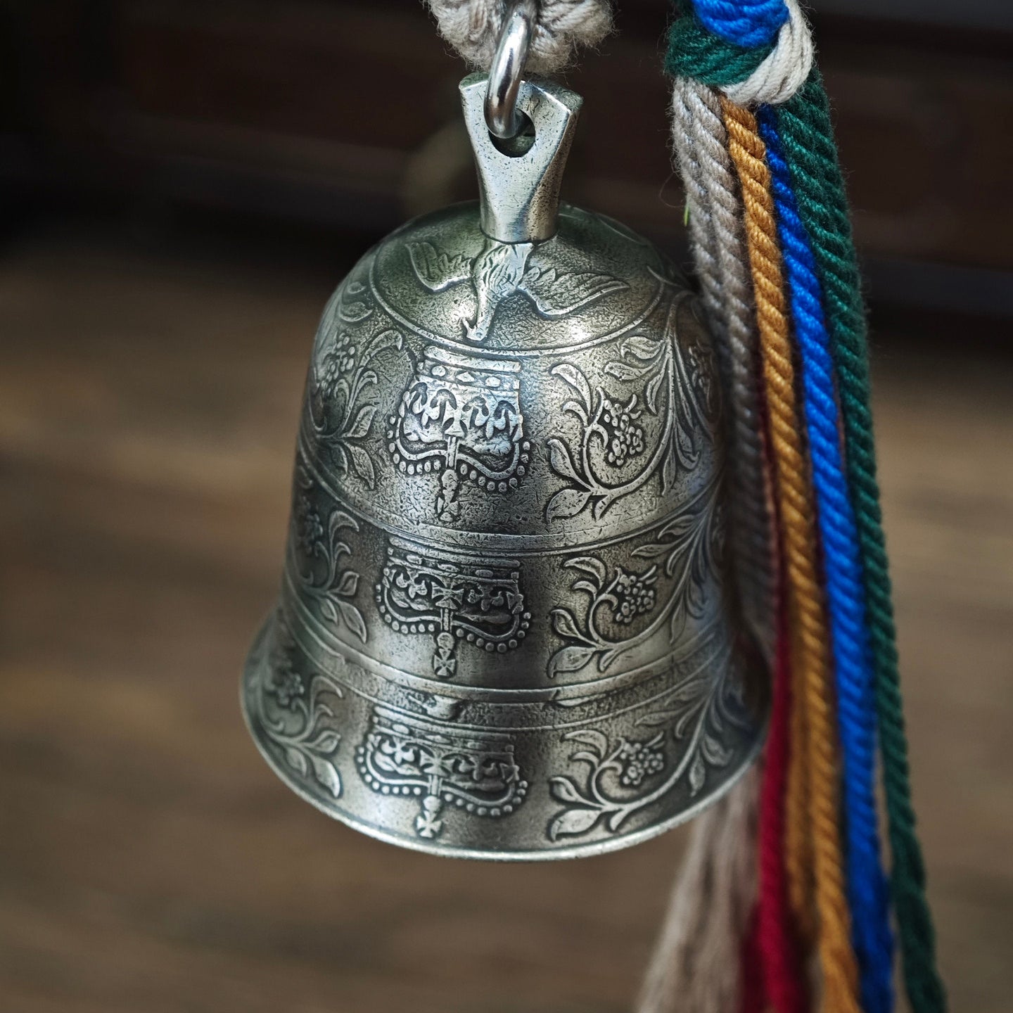 "An ornate, antiqued silver-toned metal bell pendant, featuring three horizontal bands of intricate, traditional hand-engraved floral scrollwork and decorative motifs across its curved surface. It is suspended from a polished silver-toned metal bail and jump ring, attached to a thick, woven natural fiber cord. To the right, a vibrant, multicolored tassel of braided cords (combining beige, green, blue, red, and yellow threads) is secured with a woven fiber wrap at the top. The entire piece hangs against a wa