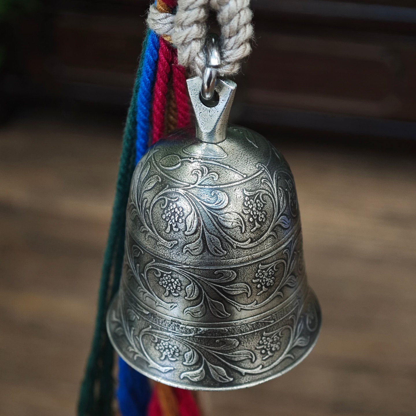 "An ornate, antiqued silver-toned metal bell pendant, featuring three horizontal bands of intricate, traditional hand-engraved floral scrollwork and decorative motifs across its curved surface. It is suspended from a polished silver-toned metal bail and jump ring, attached to a thick, woven natural fiber cord. To the right, a vibrant, multicolored tassel of braided cords (combining beige, green, blue, red, and yellow threads) is secured with a woven fiber wrap at the top. The entire piece hangs against a wa