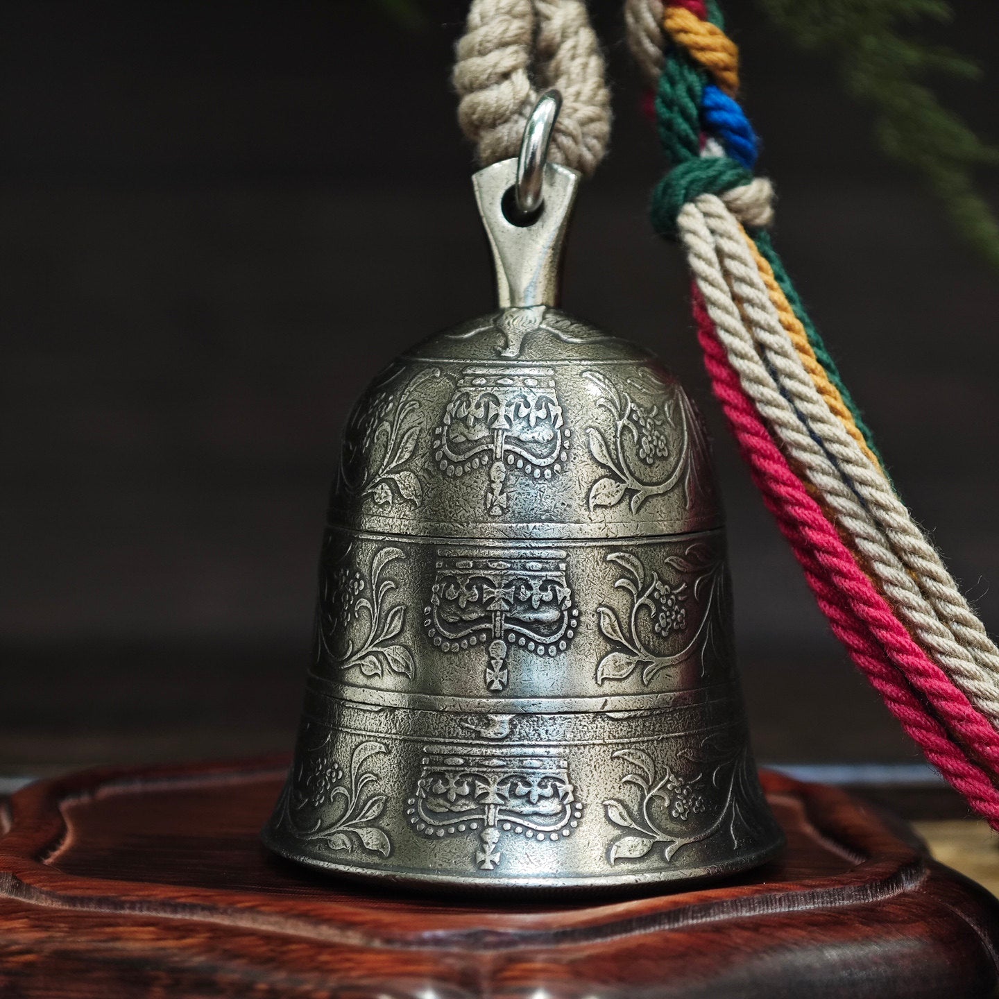 "An ornate, antiqued silver-toned metal bell, featuring three horizontal bands of intricate, hand-engraved traditional motifs and floral scrollwork across its curved surface. It is suspended from a polished silver-toned metal bail and jump ring, attached to a thick, woven natural fiber cord. To the right, a vibrant, multicolored braided tassel (combining beige, red, green, blue, and yellow threads) is secured with a woven fiber wrap. The bell rests on a carved, dark wooden base, set against a moody, blurred