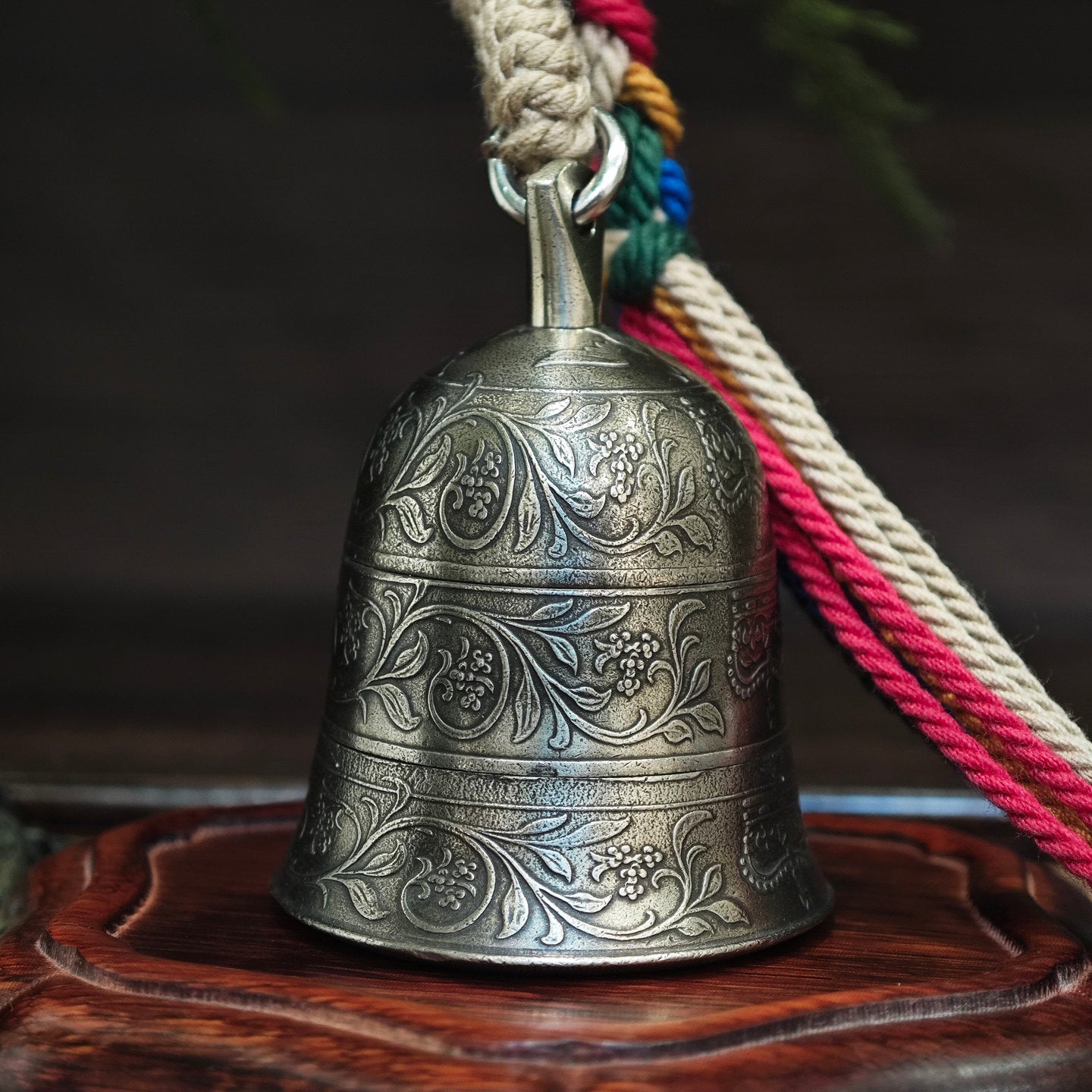"An ornate, antiqued silver-toned metal bell, featuring three horizontal bands of intricate, hand-engraved traditional motifs and floral scrollwork across its curved surface. It is suspended from a polished silver-toned metal bail and jump ring, attached to a thick, woven natural fiber cord. To the right, a vibrant, multicolored braided tassel (combining beige, red, green, blue, and yellow threads) is secured with a woven fiber wrap. The bell rests on a carved, dark wooden base, set against a moody, blurred
