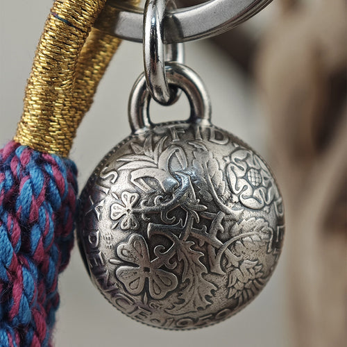 "Close-up of an ornate, antiqued silver spherical pendant with intricate hand-engraved floral and foliate patterns. It hangs from a polished silver split ring attached to a keychain, paired with a braided gold cord and a multicolored woven textile cord (pink, blue, purple) against a soft, neutral background."