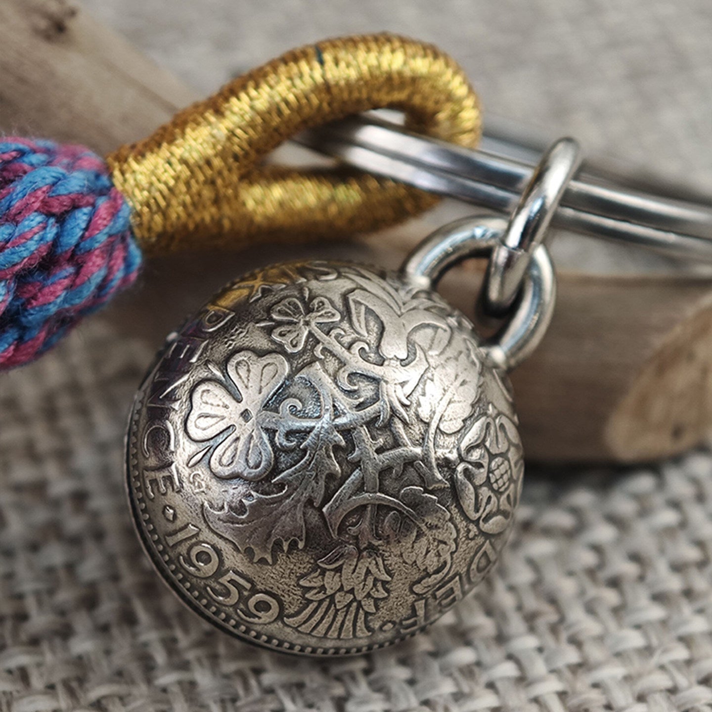 "Close-up of an antiqued silver spherical pendant, intricately engraved with floral and foliate patterns and marked with the year '1959'. It hangs from polished silver split rings attached to a keychain, paired with a braided gold cord and a vibrant multicolored woven tassel (pink, blue). The piece rests on a textured beige fabric surface with a rustic wooden accent in the background."