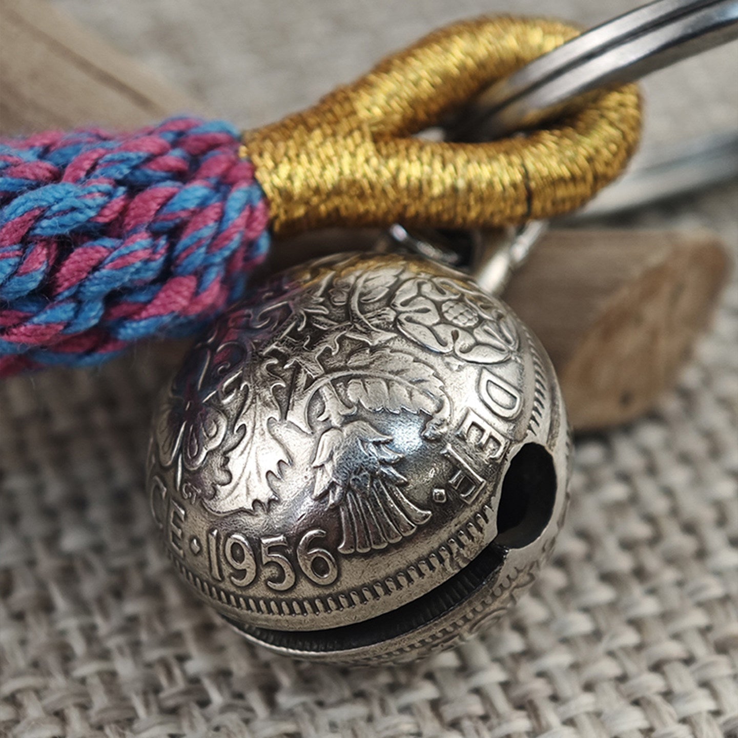 "Close-up of a vintage-style silver spherical bell pendant, featuring intricate floral engravings, the year '1956', and partial text 'DEI' across its antiqued surface. The bell has a small sound hole and hangs from polished silver split rings, paired with a braided gold cord and a vibrant pink-and-blue woven tassel, resting on a textured beige linen surface with a rustic wooden accent in the background."