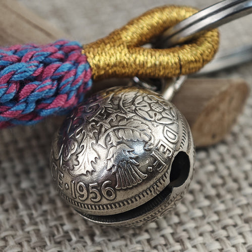 "Close-up of a vintage-style silver spherical bell pendant, featuring intricate floral engravings, the year '1956', and partial text 'DEI' across its antiqued surface. The bell has a small sound hole and hangs from polished silver split rings, paired with a braided gold cord and a vibrant pink-and-blue woven tassel, resting on a textured beige linen surface with a rustic wooden accent in the background."
