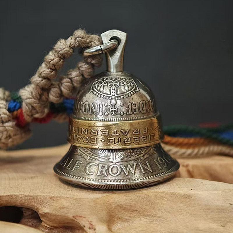"A vintage-style bell pendant crafted from repurposed coins, featuring an antiqued silver-toned body engraved with 'HALF CROWN' and a brass center band inscribed with 'LIBERTE · EGALITE · FRATERNITE'. It hangs from a braided natural jute cord with vibrant multicolored tassels, resting on a rustic live-edge wooden surface against a muted gray background."
