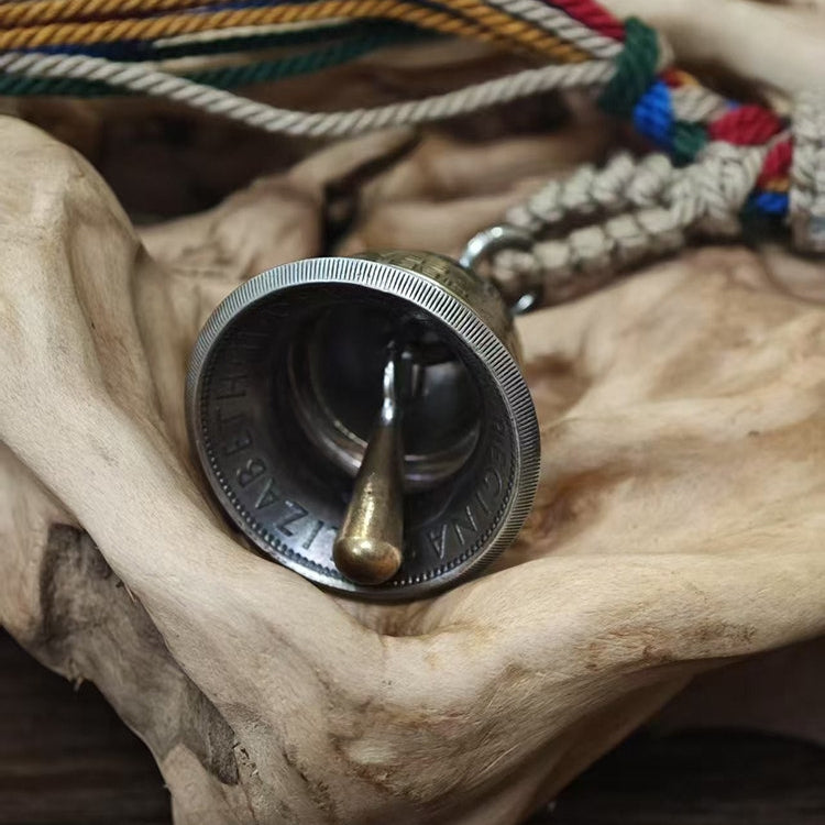 "Close-up of a repurposed silver coin bell, displayed on its side to reveal its brass clapper suspended inside. The bell features a reeded edge and partial engraved text 'ELIZABETH', resting in a natural hollow of weathered driftwood. A braided jute cord with vibrant multicolored tassels is visible in the background, all set on a rustic wooden surface."