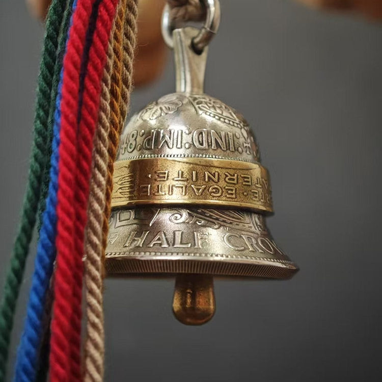 "A vintage-style bell pendant crafted from repurposed coins, featuring an antiqued silver-toned body engraved with 'HALF CROWN' and 'IND: IMP' text, accented by a polished brass center band inscribed with 'LIBERTE · EGALITE · FRATERNITE'. It hangs from a polished silver bail, with vibrant multicolored braided tassels (red, blue, green, tan, orange) to the left, set against a muted gray background."