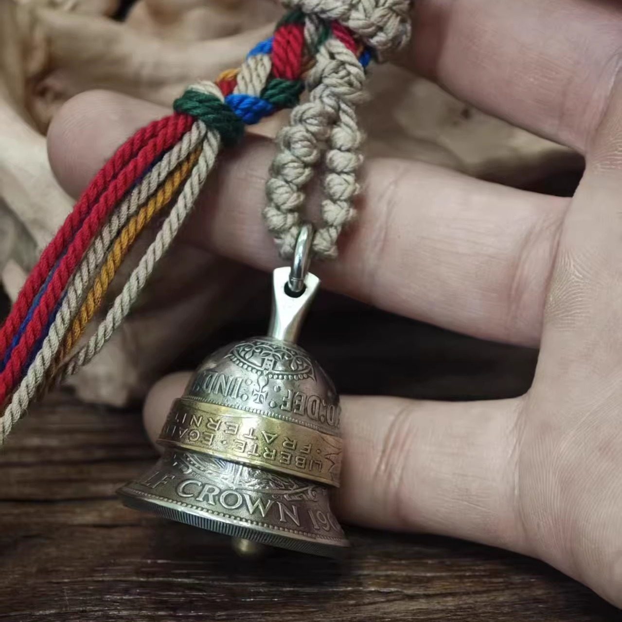 "A hand holding a vintage-style bell pendant crafted from repurposed coins, featuring an antiqued silver-toned body engraved with 'HALF CROWN' and 'IND: IMP' text, accented by a polished brass center band inscribed with 'LIBERTE · EGALITE · FRATERNITE'. It hangs from a braided natural jute cord with vibrant multicolored tassels (red, blue, green, yellow), resting against a rustic wooden surface and crumpled fabric background."