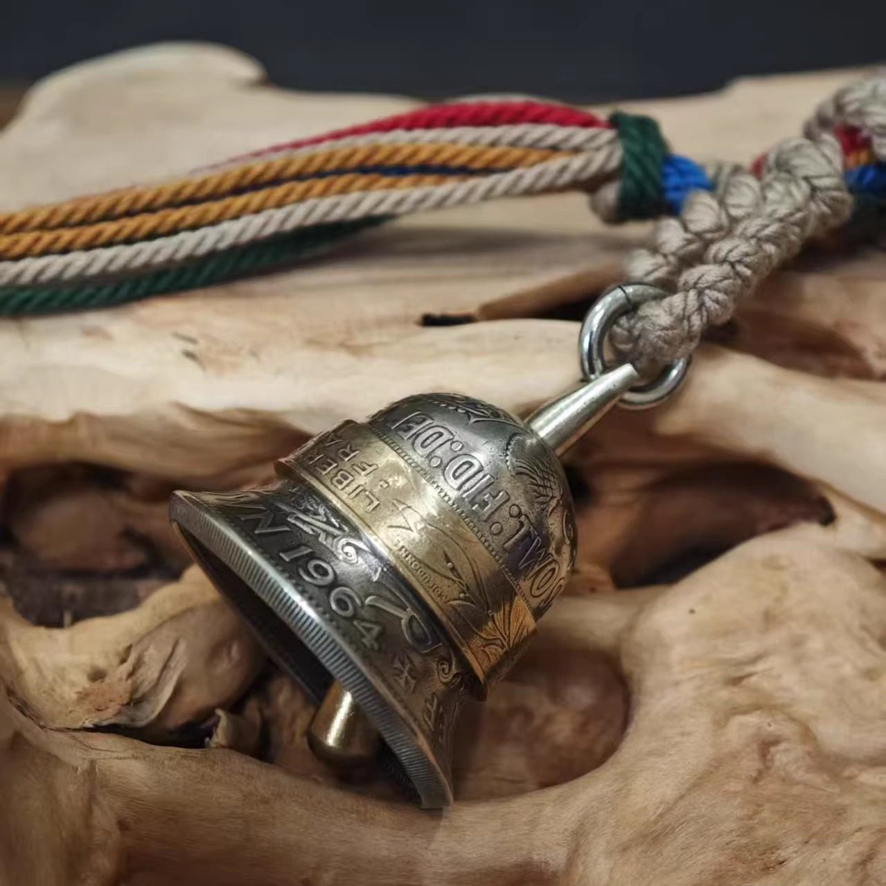 "A handcrafted vintage-style bell pendant made from repurposed coins, featuring an antiqued silver-toned body engraved with the year '1964' and partial text 'TWO', accented by a brass center band. It hangs from a braided natural jute cord with vibrant multicolored tassels (red, blue, green, yellow, tan), resting in the hollow of a weathered, live-edge driftwood surface against a muted dark background."