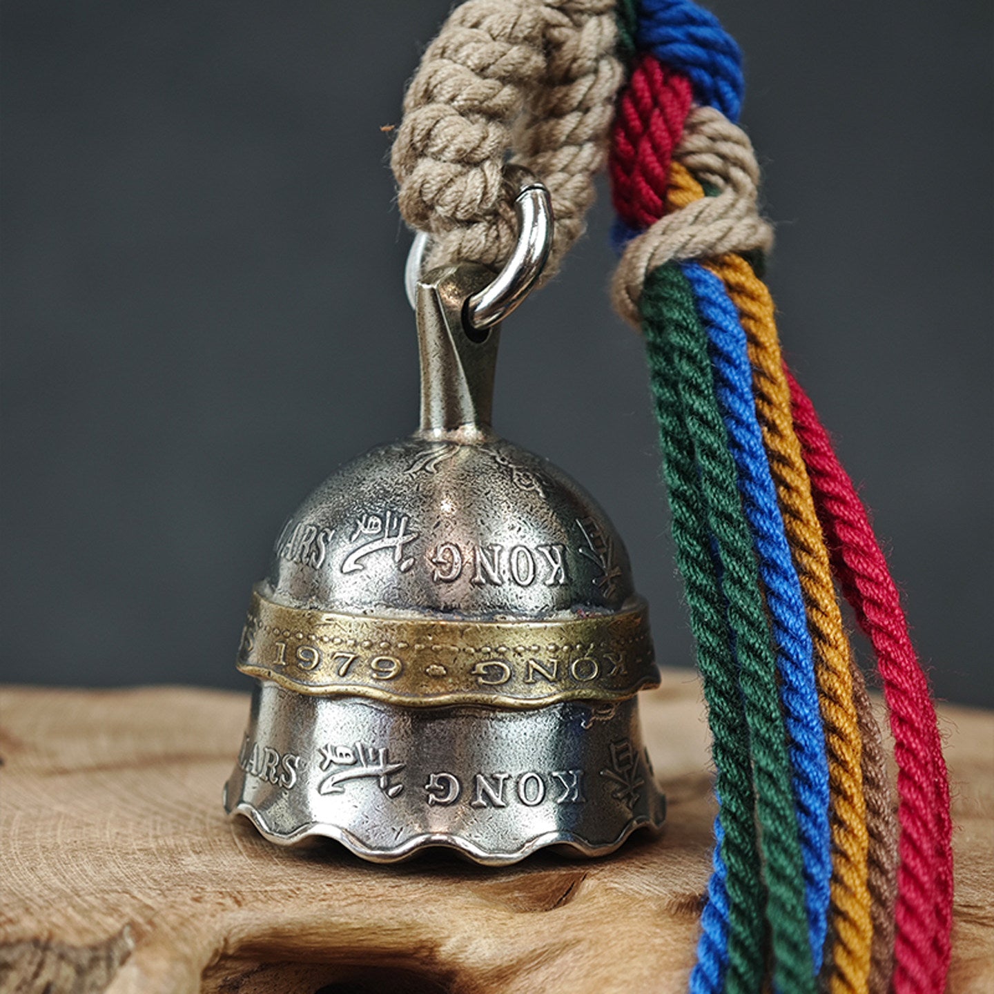 "A vintage-style metal bell with a silver-toned body, accented by a brass-colored band engraved with '1979' and 'KONG'. It is suspended from a braided jute rope hanger and paired with vibrant multicolored tassels in red, blue, green, and yellow, resting on a rustic wooden surface against a muted gray background."