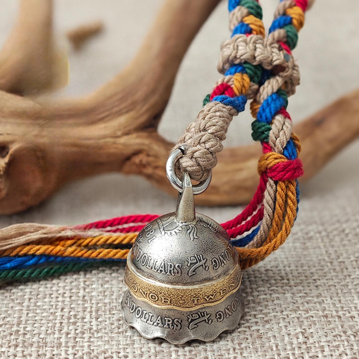 "A vintage-inspired metal bell pendant with a silver-toned body, featuring an engraved brass band that reads 'DOLLARS' and other details. It hangs from a braided natural jute cord, paired with vibrant multicolored tassels in red, blue, green, and orange, resting on a textured beige fabric surface with a rustic driftwood accent in the background."