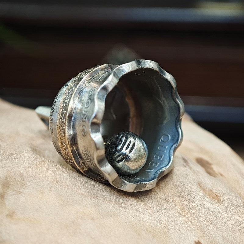 "Close-up of a vintage-style metal bell, displayed open to reveal its intricately textured metal clapper inside. The bell features a scalloped, silver-toned rim and engraved details across its exterior, resting on a smooth, light brown wooden surface against a dark, blurred background."