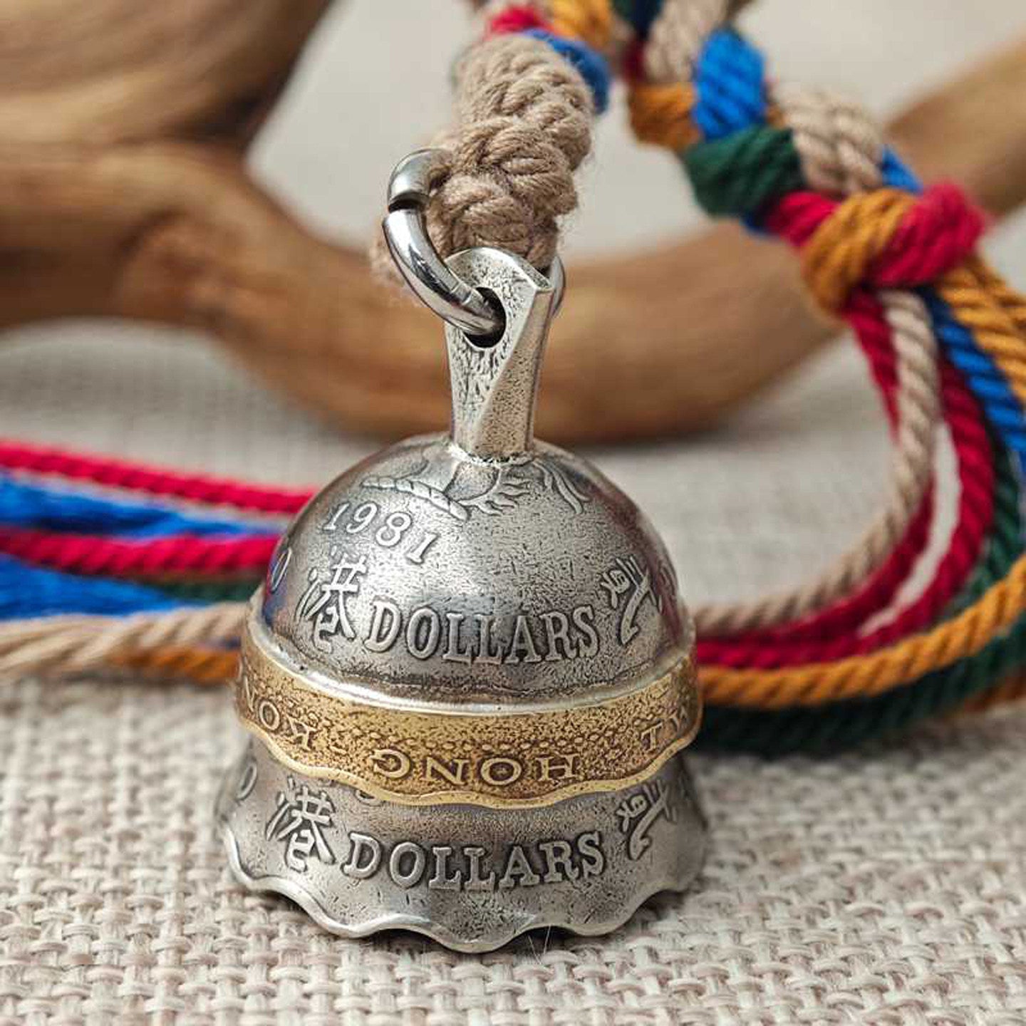 "A vintage-inspired metal bell pendant crafted to resemble repurposed Hong Kong dollar coins, featuring a silver-toned body engraved with '1981', 'HONG KONG DOLLARS', and Chinese characters, accented by a brass-colored band. It hangs from a braided natural jute cord paired with vibrant multicolored tassels in red, blue, green, and orange, resting on a textured beige fabric surface with rustic driftwood in the background."