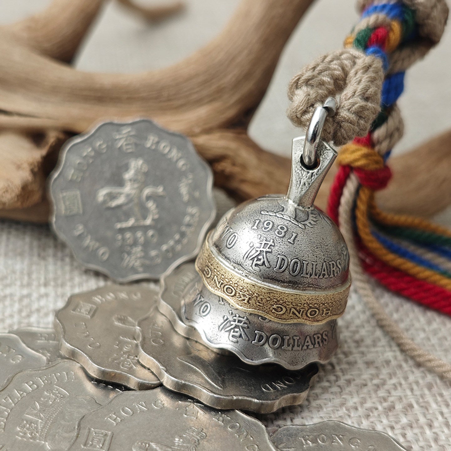 "Vintage-inspired metal bell pendant crafted from repurposed 1981 Hong Kong dollar coins, featuring a silver-toned body engraved with 'HONG KONG DOLLARS' and Chinese characters, accented by a brass-toned band. It hangs from a braided natural jute cord paired with vibrant multicolored tassels in red, blue, green, and orange, resting on a textured beige fabric surface with rustic driftwood in the background."